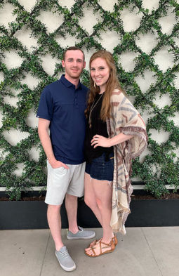 Krystina Middleton standing alongside her husband in front of a wall outlined with faux grass