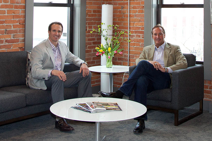 Drew Brees and John Rotche sitting in dark chairs in an office smiling. They are both dressed professional in suit coats. The wall behind them is brick with two windows. 