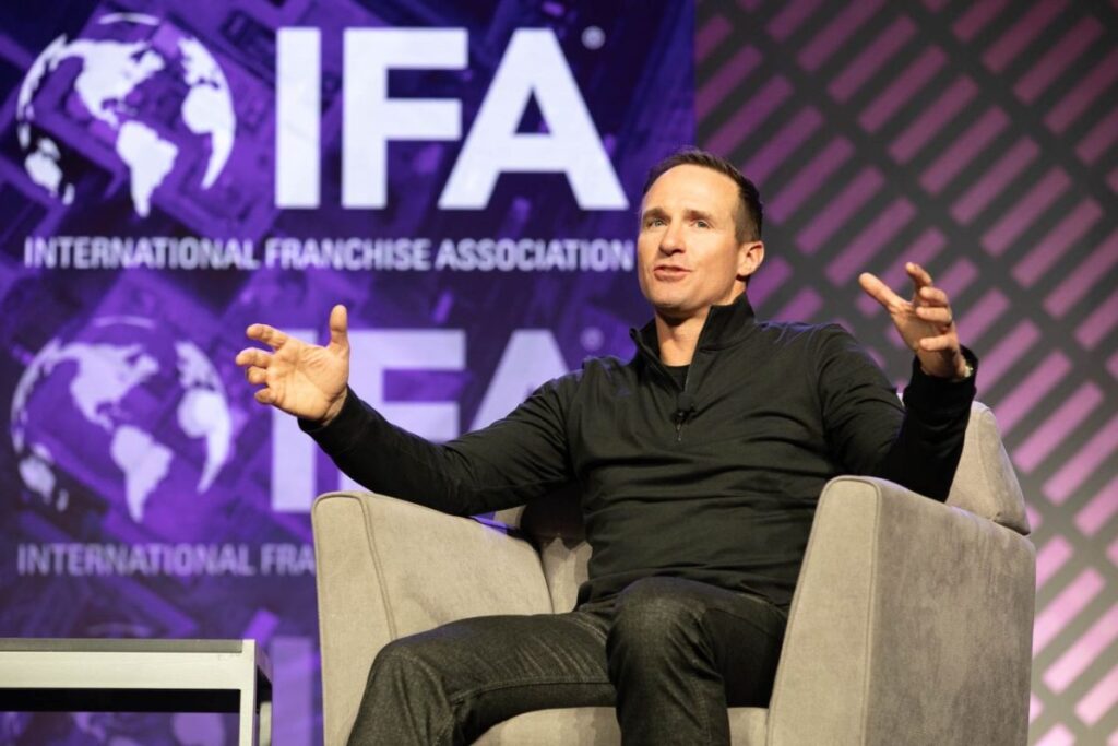 Drew Brees is sitting in a chair on stage at the IFA convention speaking to the crowd. He is in a gray chair and the screen behind him is purple with IDA graphics.