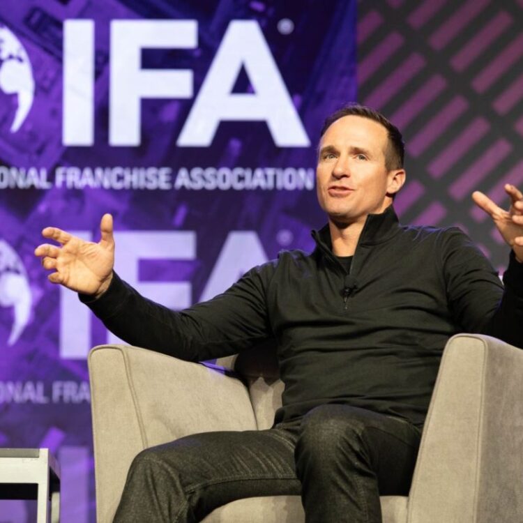 Drew Brees is sitting in a chair on stage at the IFA convention speaking to the crowd. He is in a gray chair and the screen behind him is purple with IDA graphics.