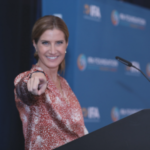 Meg Roberts is standing at a podium pointing at the camera and smiling, there is a blue vinyl banner behind her with IFA logos