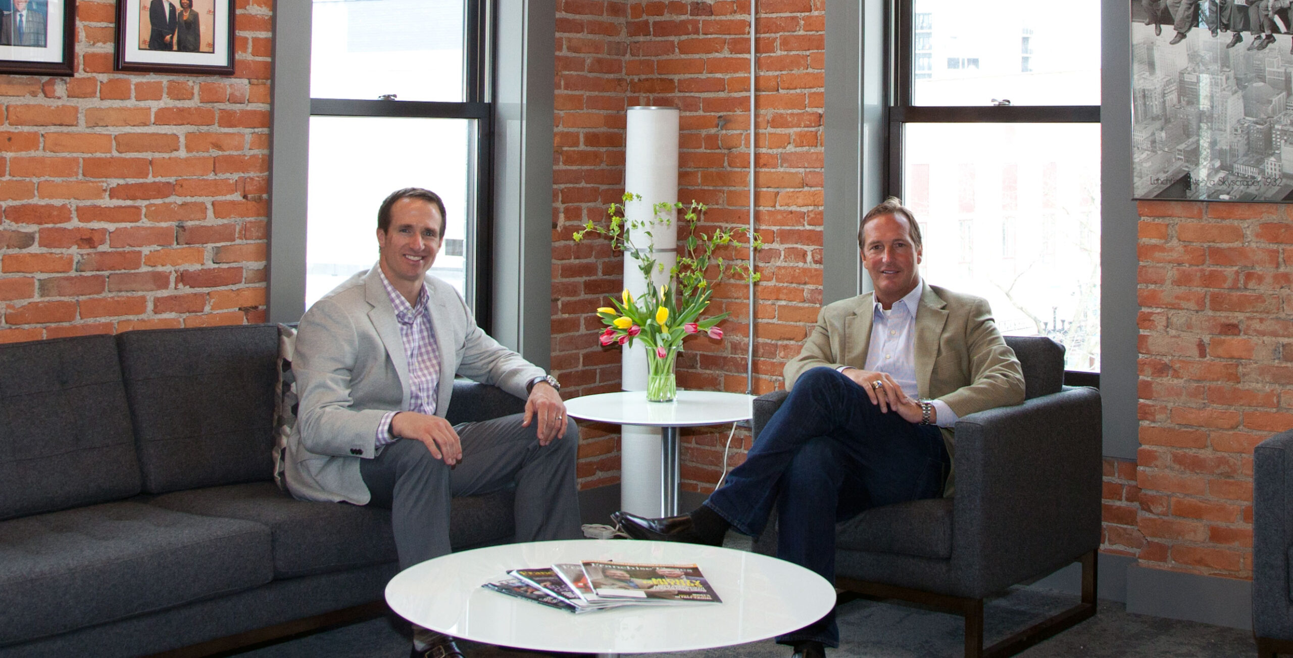 Drew Brees and John Rotche sitting in dark chairs in an office smiling. They are both dressed professional in suit coats. The wall behind them is brick with two windows. 