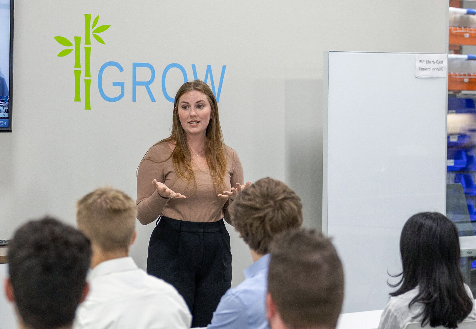 Krystina is standing and speaking to a group of interns in front of a TV, a logo that says "GROW" and a glass wall that looks into the Liberty Fulfillment Center
